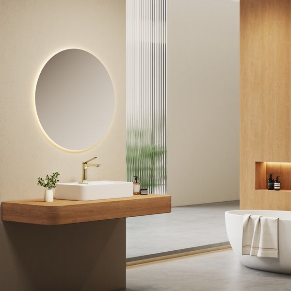 Modernes Badezimmer mit rundem, beleuchtetem Spiegel, weißem Aufsatzwaschbecken auf einer Holzkonsole, goldener Armatur und Badewanne.