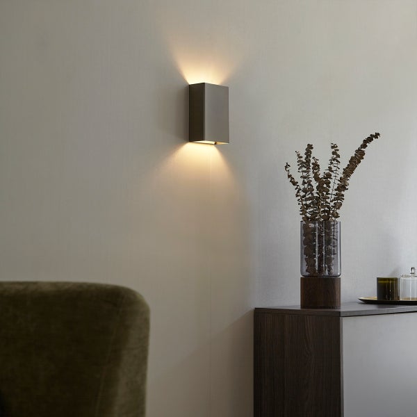 Szene mit einer grauen Wandleuchte, die ein warmes Licht ausstrahlt, und einer Vase mit Trockenblumen auf einer Kommode