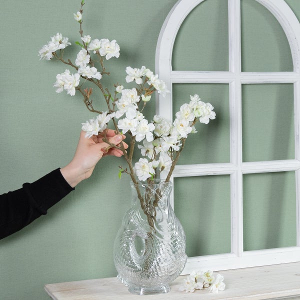 Künstliche weiße Blütenzweige in einer Glasvase auf einem hellen Holzregal vor einem weißen Fensterrahmen. Helle, organisierte Wohndekoration.