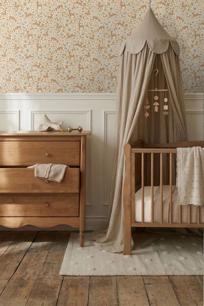 Ein Kinderzimmer mit Holzmöbeln, einem Gitterbett mit Betthimmel, einer Kommode und einer Tapete mit Tier und Blumenmuster in natürlicher Optik.