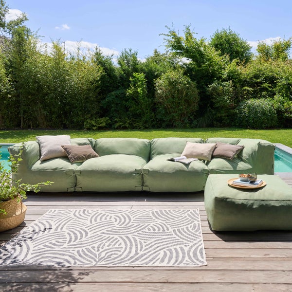 Modernes Outdoor-Lounge-Set auf einer Holzterrasse mit modularem grünem Sofa, Kissen, gemustertem Teppich und Hocker vor einem grünen Garten.