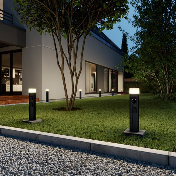 Moderner Garten mit schwarzen Pollerleuchten inklusive integrierter Steckdosen auf einer Rasenfläche neben einem Haus bei Dämmerung.