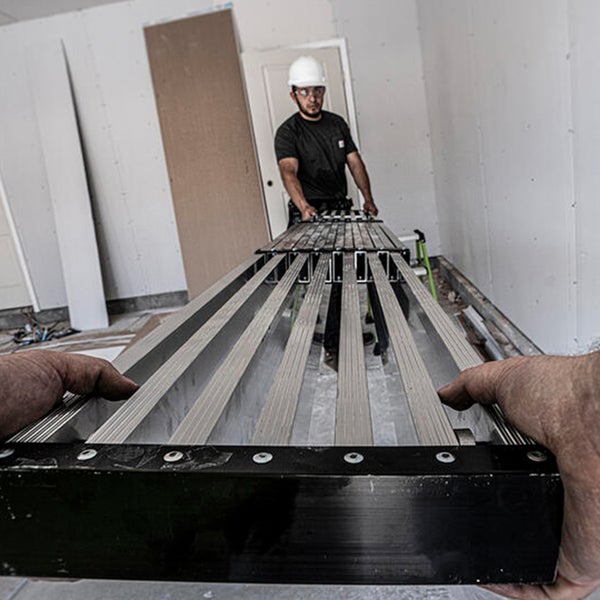Handwerker mit Schutzhelm trägt zusammen mit einer weiteren Person ein langes, ausziehbares Aluminium Gerüstteil auf einer Baustelle.