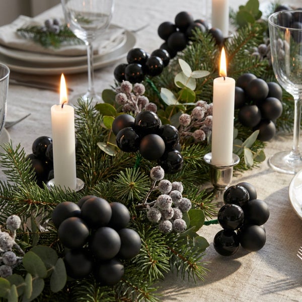 Festliche Tischdekoration mit Tannenzweigen, brennenden Stabkerzen, Eukalyptus und dekorativen Beeren auf einer gedeckten Tafel.