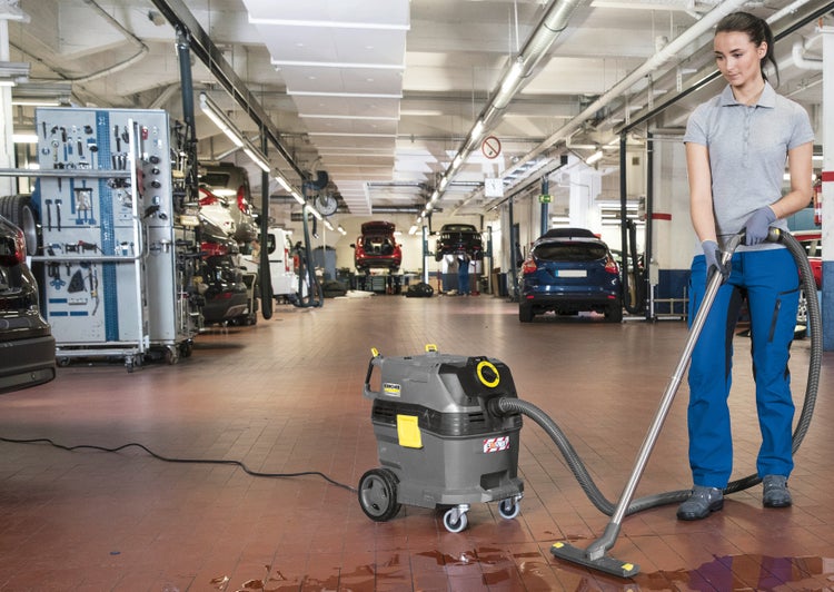Eine Frau reinigt den hellen Fliesenboden einer organisierten Fahrzeugwerkstatt mit einem Kärcher Nass- und Trockensauger.