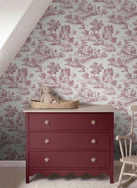 Kinderzimmer mit Toile-de-Jouy-Vliestapete, dunkelroter Kommode, Weidenkorb mit Stofftier und hellem Schaukelstuhl.