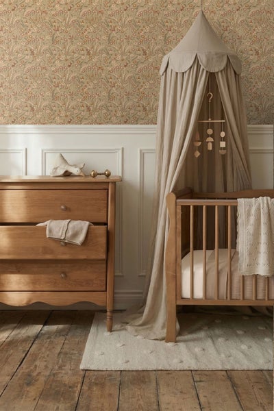 Kinderzimmer mit floraler Tapete, weißer Wandtäfelung, einer Holzkommode und einem Babybett aus Holz mit hellem Stoffbaldachin.