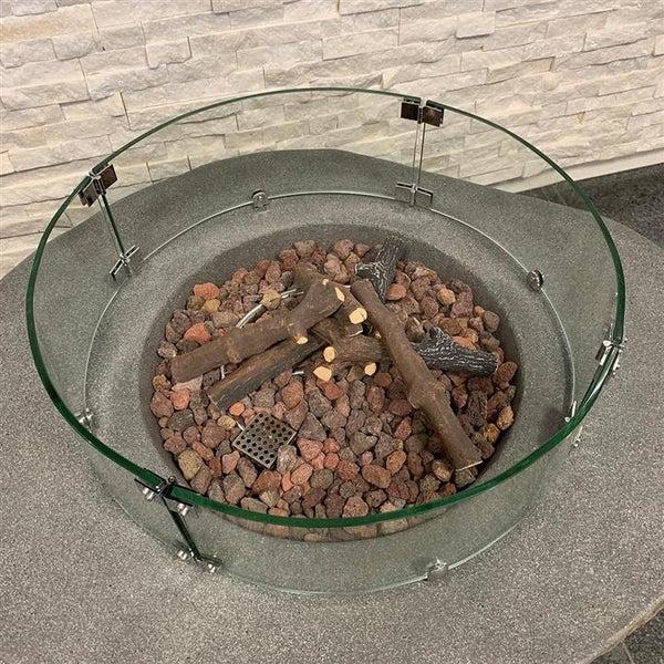 Runder Windschutz aus Glas für eine Feuerstelle mit Lavasteinen und dekorativen Holzscheiten auf einem Steintisch.