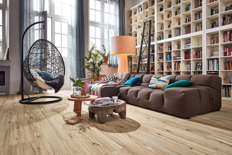 Geräumiges Wohnzimmer mit Laminatboden in Holzoptik, Hängesessel, Sofa und großem Bücherregal vor hohen Fenstern.