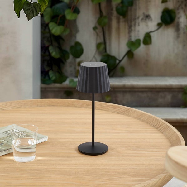 Moderne schwarze kabellose LED-Tischleuchte mit geripptem Schirm auf einem runden Holztisch im Außenbereich mit Kletterpflanzen.