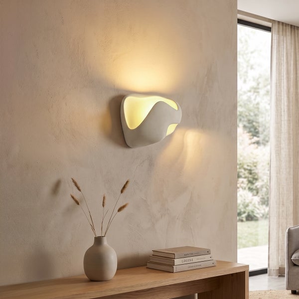 Moderne Wandleuchte mit organisch geformtem Korpus leuchtet indirekt an einer texturierten Wand über einem Sideboard mit Dekoration.