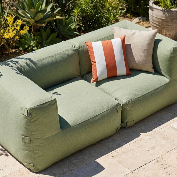 Zwei grüne gepolsterte Outdoor-Loungesessel auf einer Steinterrasse mit einem kleinen Steintisch und verschiedenen Gartenpflanzen im Hintergrund.