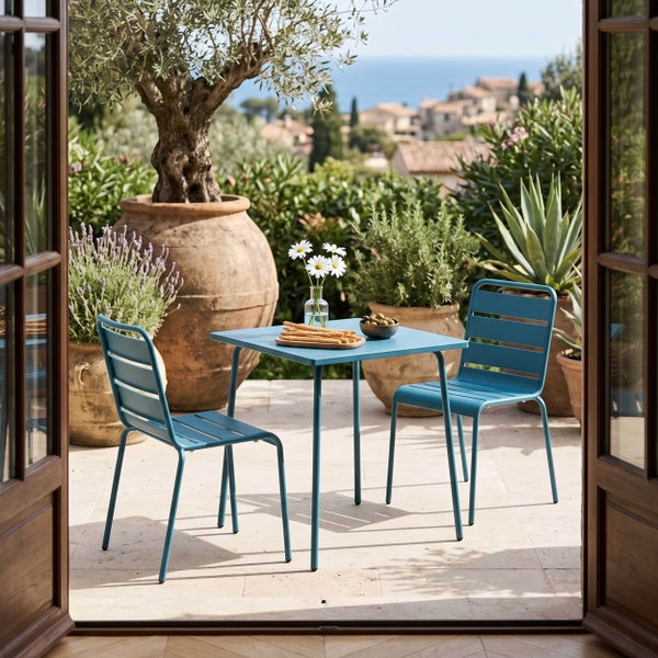 Blaues Gartenmöbel-Set aus Metall mit quadratischem Tisch und zwei Stühlen auf einer hellen Terrasse mit mediterraner Aussicht.