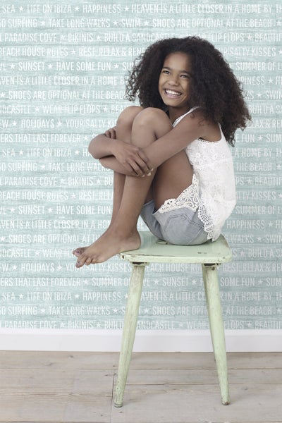 Ein lächelndes Mädchen mit lockigem Haar sitzt auf einem Hocker vor einer mit Strandmotiven bedruckten Wand.