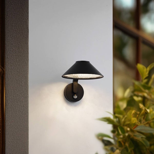 Moderne schwarze Außenwandleuchte mit Bewegungsmelder, montiert an einer weißen Wand, strahlt Licht nach unten ab.