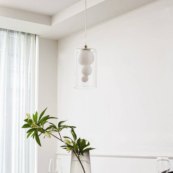 Moderne Pendelleuchte mit klarem zylindrischem Glasschirm und drei weißen Glaskugeln im Inneren, aufgehängt in einem hellen Raum mit Pflanze.