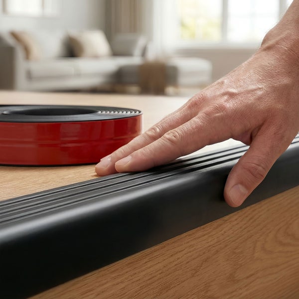 Hand drückt ein Gummiprofil als Treppenkantenprofil auf eine Holztreppenstufe neben einer aufgerollten Rolle des Profils.