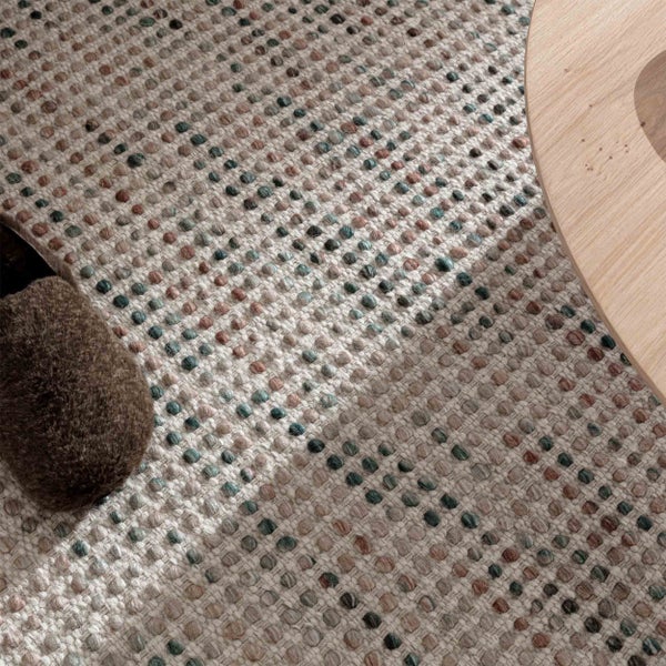 Nahaufnahme eines gewebten Teppichs mit Noppenstruktur und mehrfarbigem Punktmuster, neben einem Holztisch und einem flauschigen Hausschuh.