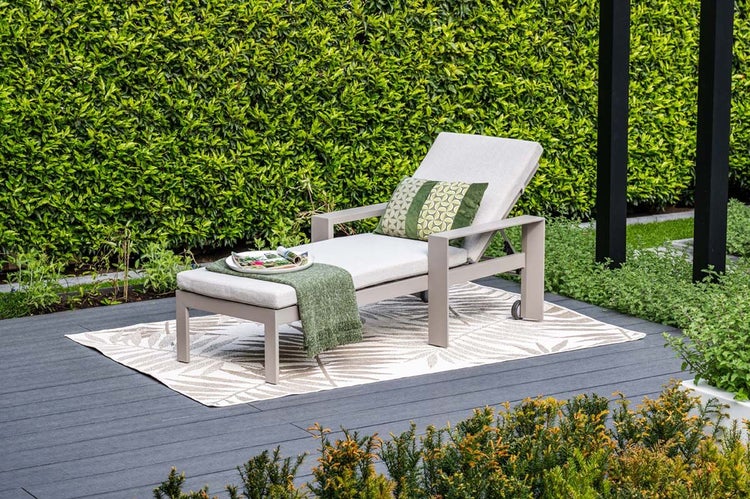 Moderne Gartenliege mit hellgrauen Polstern, Armlehnen, grüner Decke und Kissen auf einer Terrasse mit Outdoor-Teppich vor einer grünen Hecke.