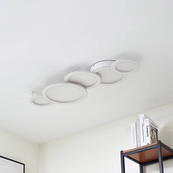 Moderne weiße Deckenleuchte mit fünf überlappenden runden Paneelen an einer hellen Zimmerdecke über einem Regal mit Büchern.