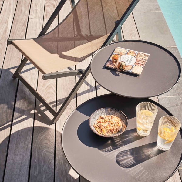 Moderne Außenszene auf Holzterrasse mit klappbarem Metall-Liegestuhl und zwei runden Beistelltischen mit Snacks, Getränken und einer Zeitschrift.