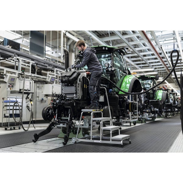 Ein Monteur nutzt eine robuste Montage-Trittstufe aus Aluminium in einer industriellen Fertigungshalle bei der Montage eines Traktors.