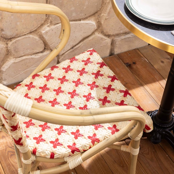 Bistro-Stuhl mit geflochtener Sitzfläche in Rot und Weiß und Rattan-Gestell auf einer Holzterrasse neben einem Bistro-Tisch vor einer Steinwand.