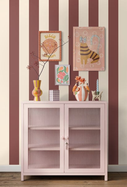 Rosa Vitrinenschrank mit geriffelten Glastüren vor einer gestreiften Tapete in Terrakotta und Creme, dekoriert mit bunten Vasen und Wandbildern.