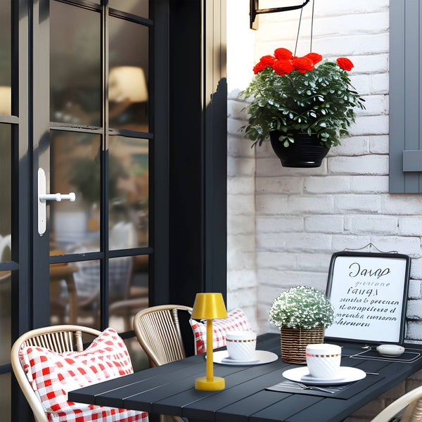 Einladender Außenbereich mit schwarzem Tisch, Stühlen mit rot-weiß karierten Kissen, gelber Tischleuchte und Blumenampel an weißer Ziegelwand.