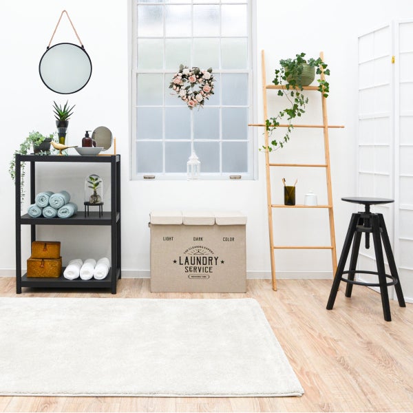 Heller, organisierter Raum mit einem großen, beigefarbenen Wäschesammler aus Stoff, einem schwarzen Regal und einer hölzernen Handtuchleiter.