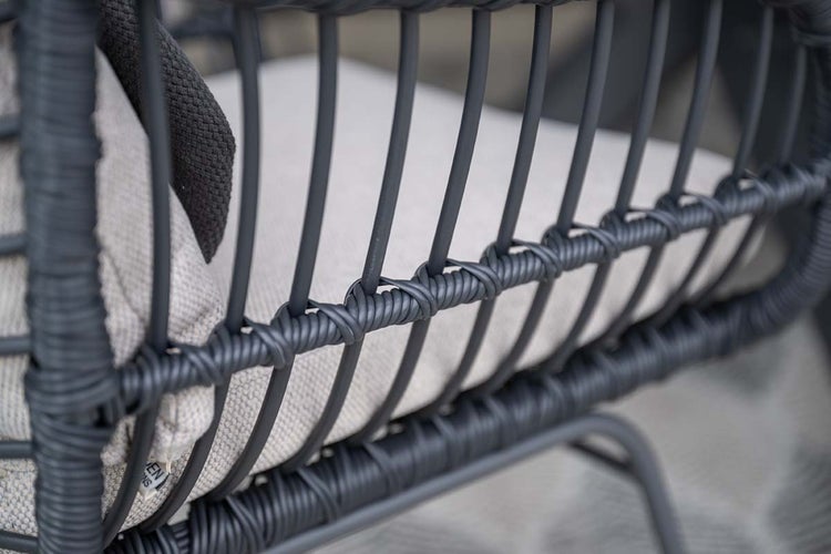 Nahaufnahme eines Loungesessels mit dunklem Geflechtrahmen, vertikalen Stäben und einem hellen Sitzkissen.