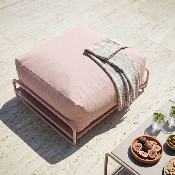 Moderner quadratischer Gartenhocker in Rosa mit Metallgestell auf einer hellen Steinterrasse neben einem Beistelltisch mit Snacks.