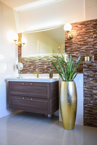 Modernes Badezimmer mit dunkelbraunem Waschtischunterschrank, goldener Armatur, großem Spiegel, braunen Mosaikfliesen und goldener Vase mit Pflanze.