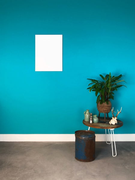 Türkis gestrichene Wand mit leerem weißem Rahmen, Beistelltisch aus Holz mit Pflanze und Deko sowie Metalltonne auf einem Betonboden.
