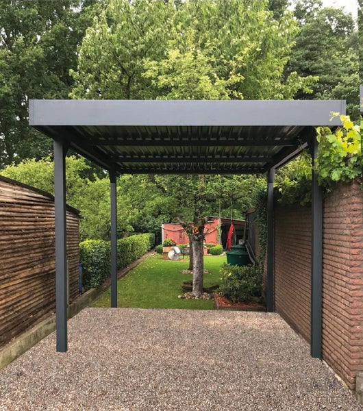 Anthrazitfarbenes Metall-Carport mit Flachdach auf einer Kiesfläche in einem hellen Garten zwischen einer Holzhütte und einer Ziegelwand.