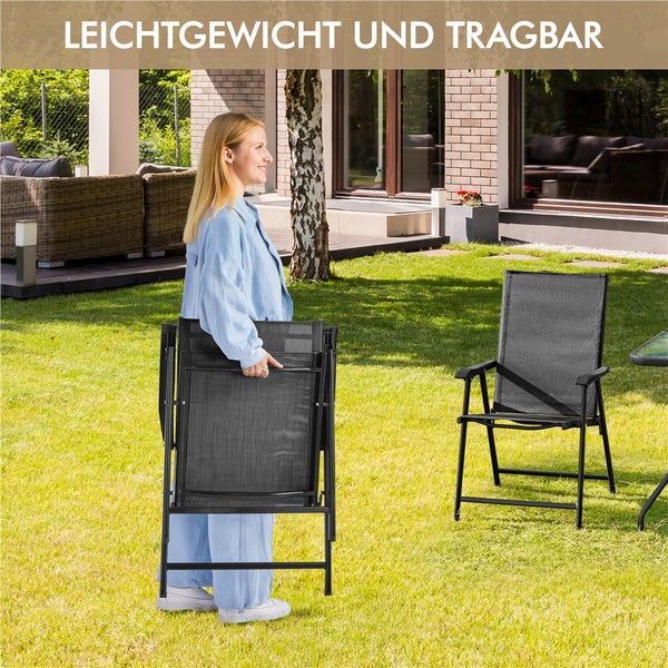 Eine Frau trägt einen zusammengeklappten Gartenstuhl aus Metall und Textilgewebe auf einem Rasen neben einem aufgeklappten Stuhl. Leichtgewicht und tragbar.
