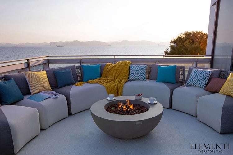 Moderne Outdoor-Lounge auf einer Terrasse mit geschwungenem Sofa, bunten Kissen und einem runden Feuertisch mit Blick auf das Meer.