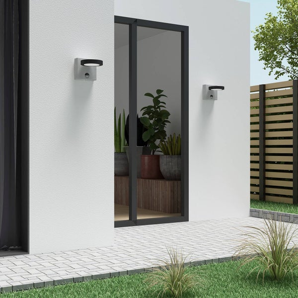 Moderne Außenwandleuchten mit Bewegungsmelder an einer weißen Fassade neben einer Terrassentür, gepflasterter Außenbereich mit Rasen.