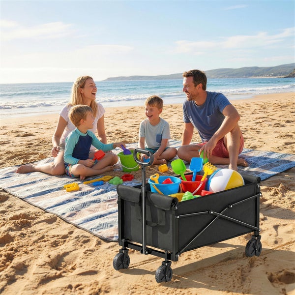 Eine glückliche Familie spielt mit einem faltbaren Bollerwagen voller Sandspielzeug am Strand.