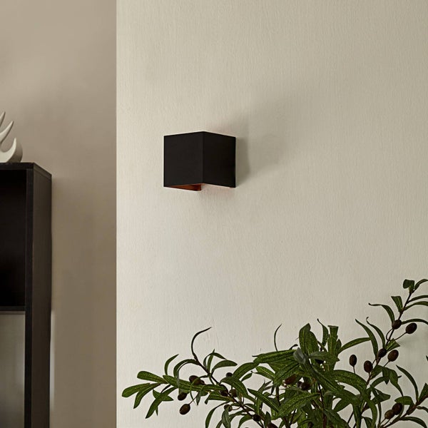 Moderne, würfelförmige Wandleuchte in Schwarz mit kupferfarbener Innenseite an einer hellen Wand neben einer Zimmerpflanze.