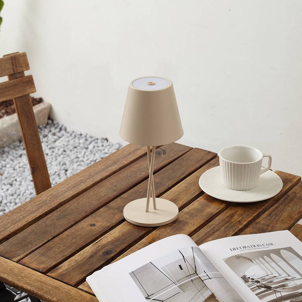 Moderne Akku-Tischleuchte auf einem Holztisch im Außenbereich neben einer Kaffeetasse und einem aufgeschlagenen Buch.