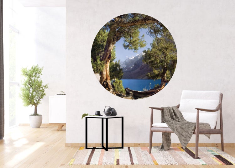 Wandbild mit einer See- und Berglandschaft im runden Rahmen in einem hellen Raum mit Sessel und Tisch