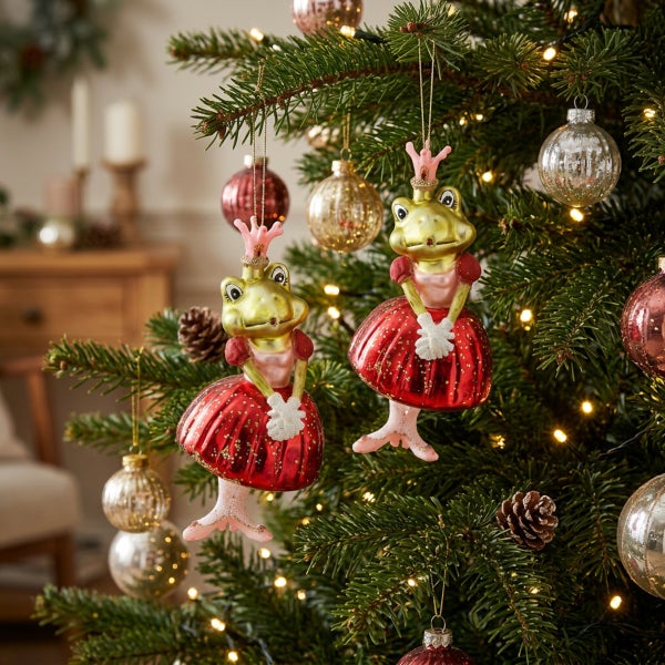 Zwei gläserne Froschprinzessin Weihnachtsanhänger mit Krone und Kleid hängen als Weihnachtsbaumschmuck in einem geschmückten Tannenbaum.