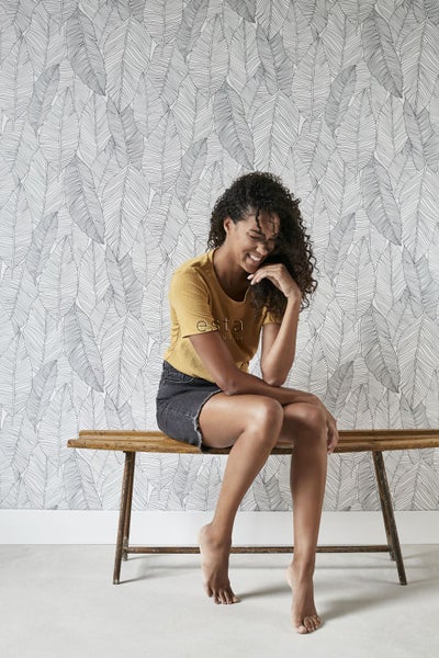 Frau sitzt vor einer Wand mit Blättertapete auf einer Holzbank.