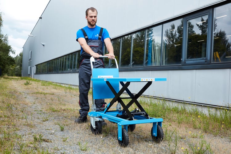 Ein Mann in Arbeitskleidung schiebt einen fahrbaren Scherenhubtisch im Außenbereich vor einem Industriegebäude auf Schotter. HanseLifter Logo.