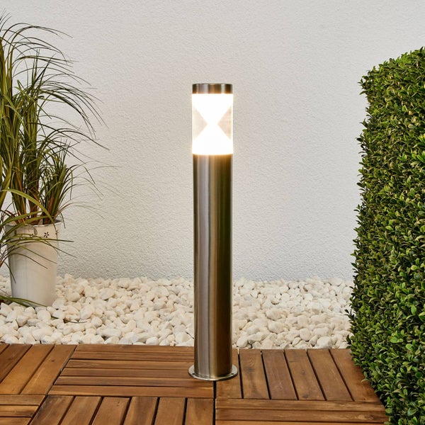 Moderne Pollerleuchte aus Edelstahl auf einer Holzterrasse neben weißem Kies und grünen Pflanzen.