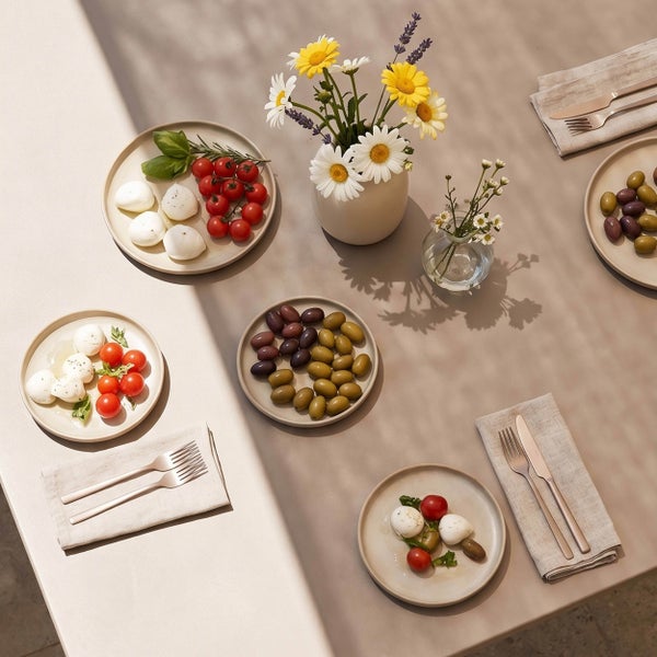 Modern gedeckter Tisch mit Keramiktellern, Mozzarella, Tomaten, Oliven, Leinenservietten, Metallbesteck und Vasen mit Blumen.