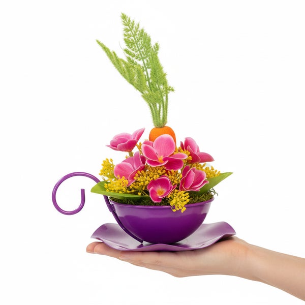 Eine Hand hält eine violette Metalldekoration in Form einer Tasse mit Untertasse, dekoriert mit künstlichen Frühlingsblumen und einer Karotte.