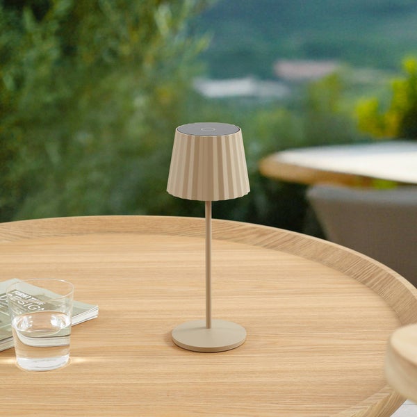 Moderne beige LED-Tischleuchte mit plissiertem Schirm auf einem runden Holztisch in einer hellen Außenumgebung.