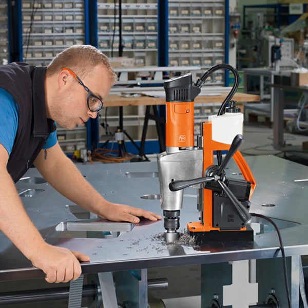 Ein Mann mit Schutzbrille bedient eine Magnet-Kernbohrmaschine, um ein Loch in eine Metallplatte in einer Werkstatt zu bohren.